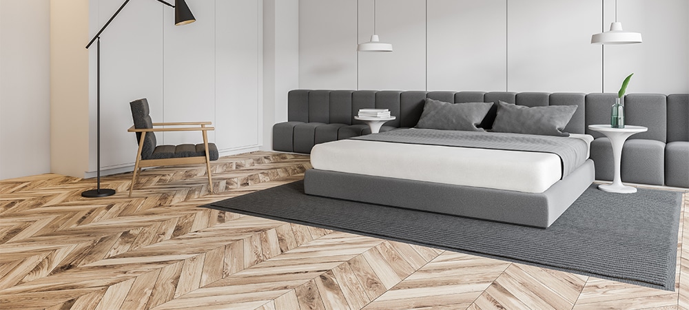 bedroom with white walls, chevron wooden floor, master bed standing on gray carpet and armchair with floor lamp near it